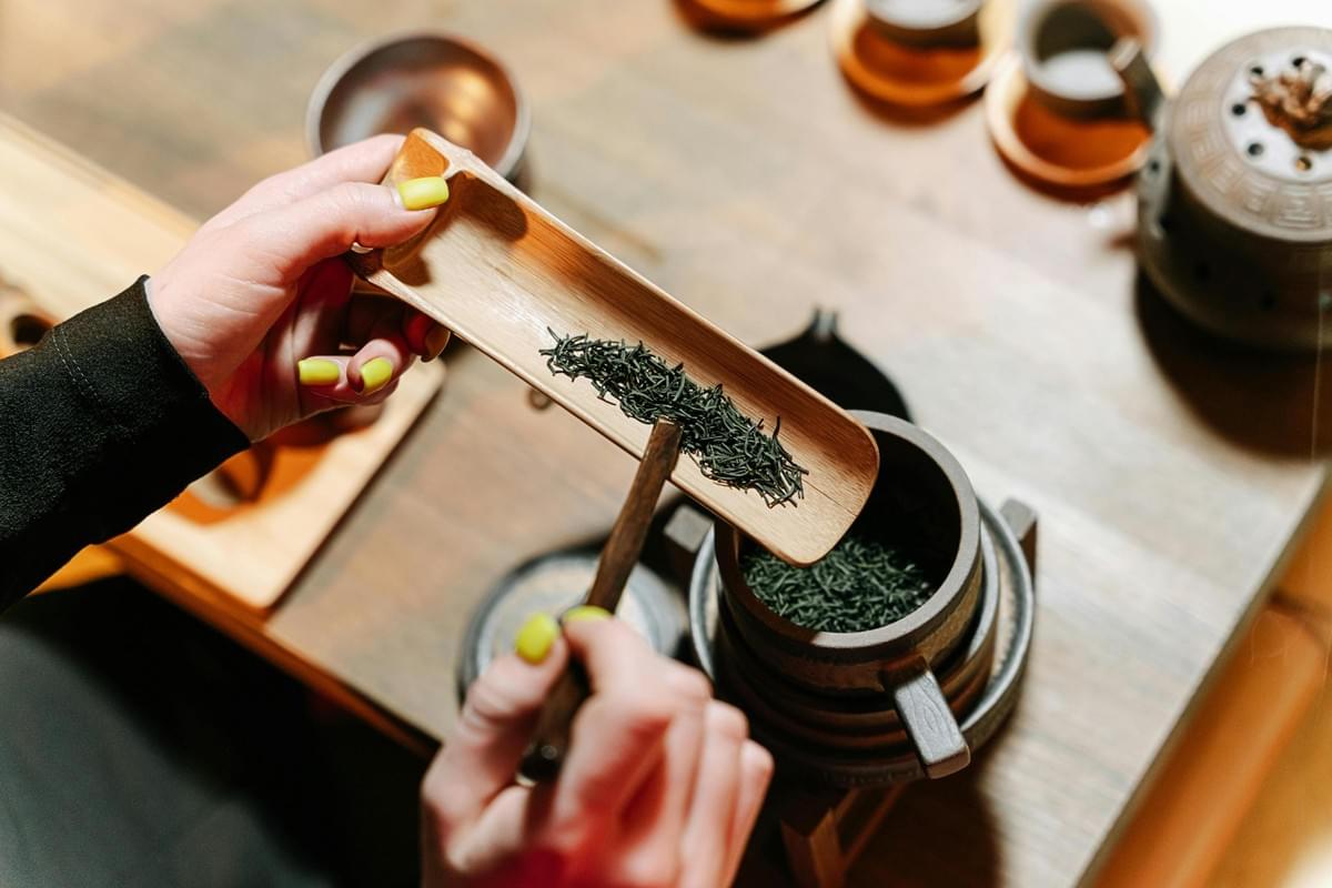 A person carefully uses a thin wooden tool to guide loose green tea leaves from a bamboo scoop into a dark, textured teapot. The close-up shot highlights the ritualistic preparation, showcasing traditional wooden accessories and ceramic teaware arranged on a wooden table. A person carefully uses a thin wooden tool to guide loose green tea leaves from a bamboo scoop into a dark, textured teapot. The close-up shot highlights the ritualistic preparation, showcasing traditional wooden accessories and ceramic teaware arranged on a wooden table.
