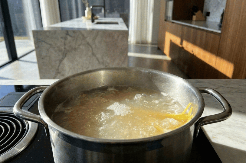 A stainless steel pot filled with boiling water and submerged spaghetti noodles on a modern induction cooktop, with a blurred marble kitchen island in the background. A stainless steel pot filled with boiling water and submerged spaghetti noodles on a modern induction cooktop, with a blurred marble kitchen island in the background.
