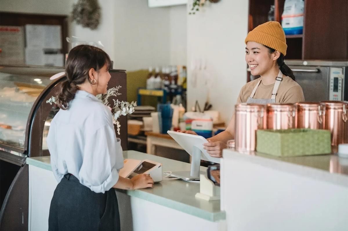Smiling barista in a yellow beanie taking an order from a customer at a cafe counter with copper canisters. Smiling barista in a yellow beanie taking an order from a customer at a cafe counter with copper canisters.