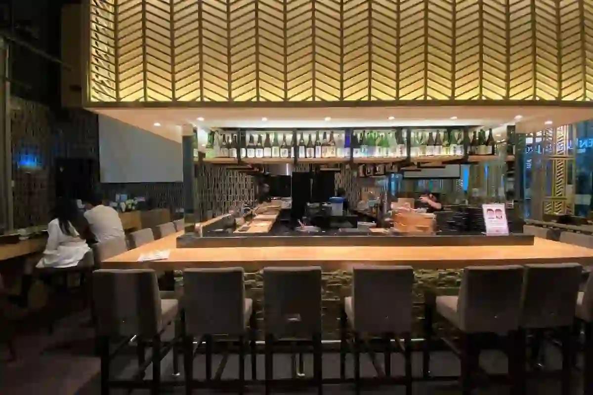 A warm, modern Japanese restaurant features an L-shaped wooden bar counter lined with several grey high-back stools. Above the bar, a large illuminated canopy with a geometric chevron pattern hangs over shelves of sake bottles and a glimpse of the kitchen staff. A warm, modern Japanese restaurant features an L-shaped wooden bar counter lined with several grey high-back stools. Above the bar, a large illuminated canopy with a geometric chevron pattern hangs over shelves of sake bottles and a glimpse of the kitchen staff.