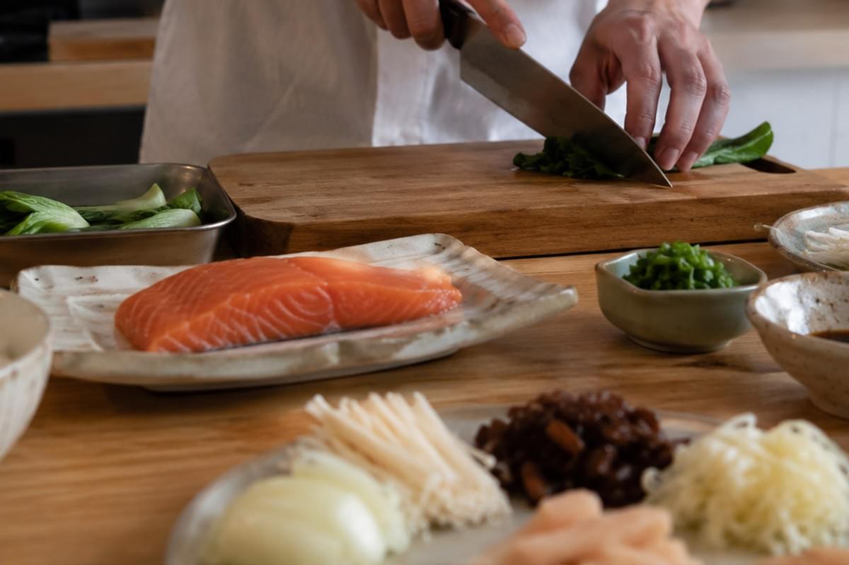 A chef dressed in a white uniform is captured from the chest down, carefully slicing fresh green leafy vegetables with a large chef's knife on a sturdy wooden cutting board. In the foreground, a vibrant, thick fillet of raw orange salmon rests on an artisanal white ceramic plate, surrounded by an organized mise en place on a smooth wooden tabletop. The culinary preparation includes a metal tray filled with bok choy leaves, a small green bowl of finely chopped scallions, a dish containing dark fermented condiments, and a large round platter displaying neatly separated piles of sliced onions, enoki mushrooms, minced ingredients, and shredded garnishes. A chef dressed in a white uniform is captured from the chest down, carefully slicing fresh green leafy vegetables with a large chef's knife on a sturdy wooden cutting board. In the foreground, a vibrant, thick fillet of raw orange salmon rests on an artisanal white ceramic plate, surrounded by an organized mise en place on a smooth wooden tabletop. The culinary preparation includes a metal tray filled with bok choy leaves, a small green bowl of finely chopped scallions, a dish containing dark fermented condiments, and a large round platter displaying neatly separated piles of sliced onions, enoki mushrooms, minced ingredients, and shredded garnishes.