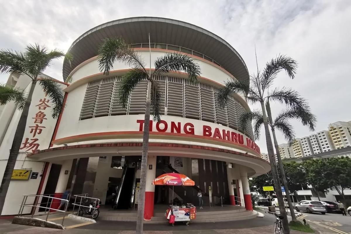 The modern, curved facade of Tiong Bahru Market stands prominently with its name displayed in bold red lettering across the white exterior. Palm trees frame the entrance, where an ice cream cart sits under a colorful umbrella on the wide sidewalk. The modern, curved facade of Tiong Bahru Market stands prominently with its name displayed in bold red lettering across the white exterior. Palm trees frame the entrance, where an ice cream cart sits under a colorful umbrella on the wide sidewalk.