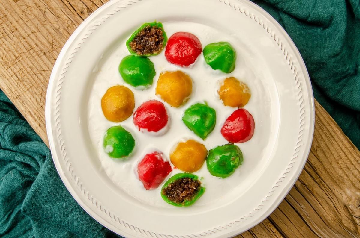 Colorful red, green, and yellow glutinous rice balls rest in a shallow white plate filled with sweet coconut milk sauce. Two of the green dumplings are bitten or split open, revealing a sweet dark coconut filling inside. Colorful red, green, and yellow glutinous rice balls rest in a shallow white plate filled with sweet coconut milk sauce. Two of the green dumplings are bitten or split open, revealing a sweet dark coconut filling inside.