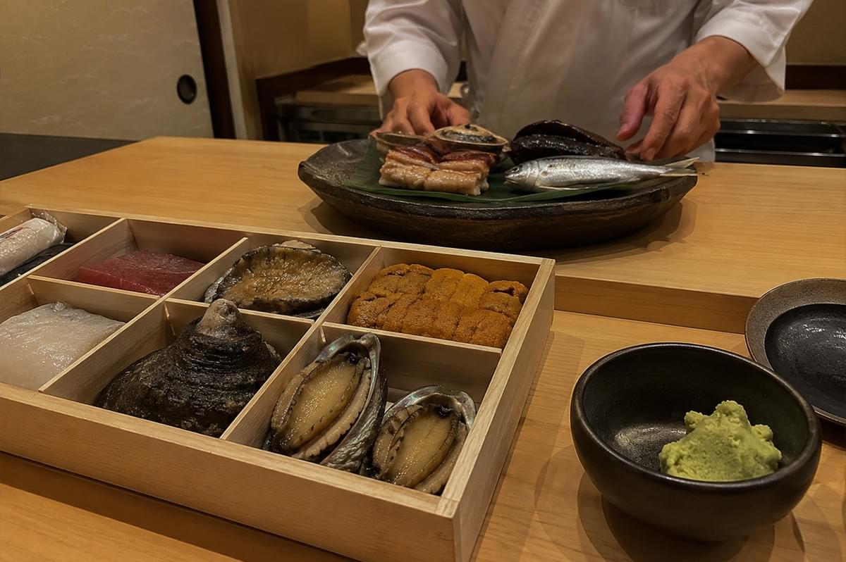 A high-angle, detailed shot captures a selection of premium sushi ingredients beautifully arranged on a light wooden counter during an omakase dining experience. In the foreground, a multi-compartment wooden box displays neatly separated delicacies, including a bright red cut of tuna, rows of vibrant orange uni (sea urchin), a textured dark abalone shell, and freshly shucked abalone glistening in their shells. To the right sits a small, dark ceramic bowl containing a generous portion of freshly grated green wasabi. In the background, under warm ambient light, a sushi chef dressed in a traditional white uniform is positioned behind a large, rustic ceramic platter lined with a green leaf, showcasing more seafood such as a whole silver fish and cooked eel, creating a refined and authentic culinary atmosphere. A high-angle, detailed shot captures a selection of premium sushi ingredients beautifully arranged on a light wooden counter during an omakase dining experience. In the foreground, a multi-compartment wooden box displays neatly separated delicacies, including a bright red cut of tuna, rows of vibrant orange uni (sea urchin), a textured dark abalone shell, and freshly shucked abalone glistening in their shells. To the right sits a small, dark ceramic bowl containing a generous portion of freshly grated green wasabi. In the background, under warm ambient light, a sushi chef dressed in a traditional white uniform is positioned behind a large, rustic ceramic platter lined with a green leaf, showcasing more seafood such as a whole silver fish and cooked eel, creating a refined and authentic culinary atmosphere.