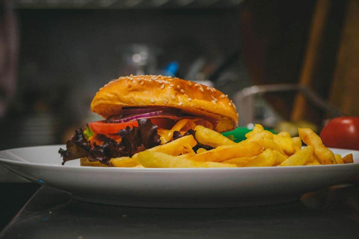 A juicy burger with a sesame seed bun, tomato, red onion, and dark leafy greens is served on a white plate alongside a heap of golden french fries. The meal is captured in a close-up shot with a softly blurred dark kitchen background. A juicy burger with a sesame seed bun, tomato, red onion, and dark leafy greens is served on a white plate alongside a heap of golden french fries. The meal is captured in a close-up shot with a softly blurred dark kitchen background.