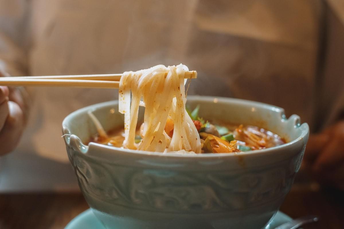 A pair of wooden chopsticks lifts a bundle of flat rice noodles from a bowl of hot, flavorful soup. The ceramic bowl is filled with a rich broth, herbs, and vegetables. A pair of wooden chopsticks lifts a bundle of flat rice noodles from a bowl of hot, flavorful soup. The ceramic bowl is filled with a rich broth, herbs, and vegetables.
