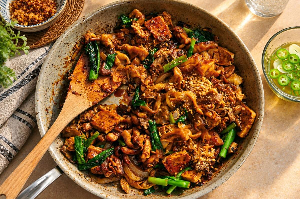 Skillet filled with stir-fried flat rice noodles, tofu, chicken, and leafy greens alongside a small dish of sliced green chilies. Skillet filled with stir-fried flat rice noodles, tofu, chicken, and leafy greens alongside a small dish of sliced green chilies.