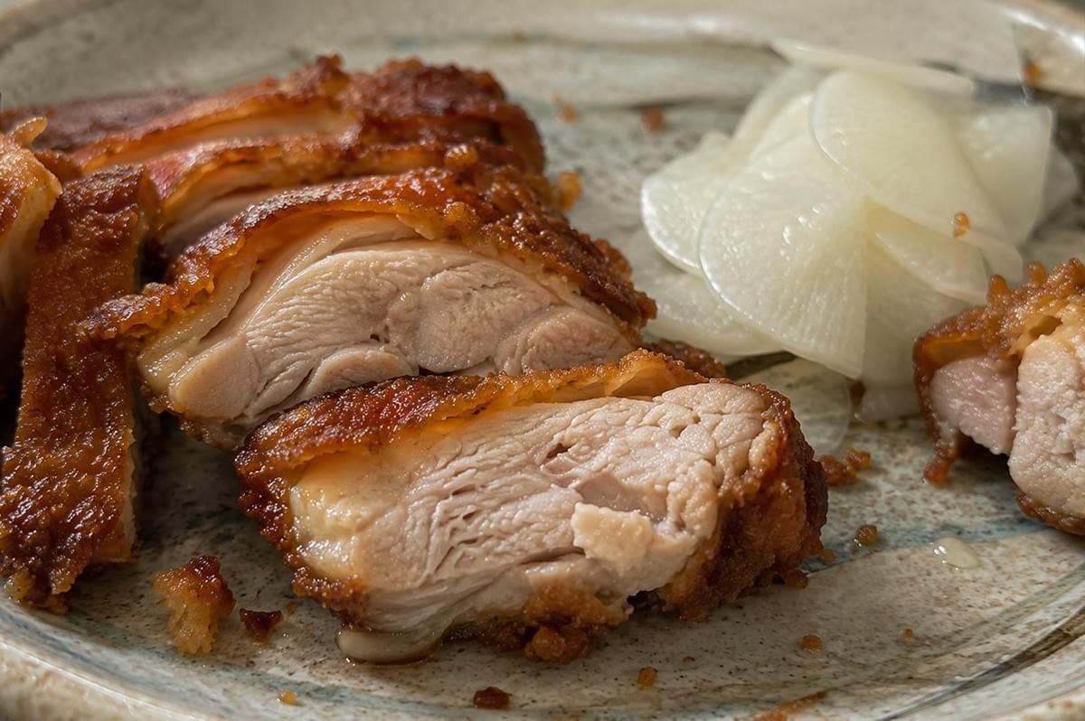 A tight, macro macro-style close-up focuses intensely on the cross-section of a freshly sliced piece of Shoyu Koji fried chicken on a rustic ceramic plate. The center slice highlights a remarkably juicy, plump, and perfectly cooked chicken thigh muscle with glistening juices, surrounded by a distinct, intensely caramelized, and jaggedly crunchy deep-amber skin. To the right, a delicate mound of translucent, paper-thin white pickled daikon radish slices provides a stark contrast to the rich, savory tones of the fried chicken. A tight, macro macro-style close-up focuses intensely on the cross-section of a freshly sliced piece of Shoyu Koji fried chicken on a rustic ceramic plate. The center slice highlights a remarkably juicy, plump, and perfectly cooked chicken thigh muscle with glistening juices, surrounded by a distinct, intensely caramelized, and jaggedly crunchy deep-amber skin. To the right, a delicate mound of translucent, paper-thin white pickled daikon radish slices provides a stark contrast to the rich, savory tones of the fried chicken.