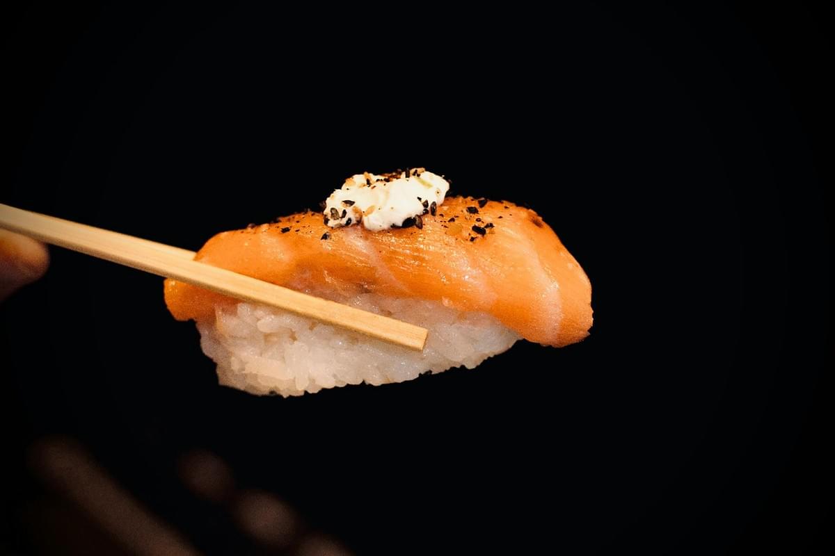 A pair of chopsticks holds a piece of salmon nigiri topped with a dollop of cream and a sprinkle of black seasoning. The vibrant orange fish and white rice stand out sharply against the solid black background. A pair of chopsticks holds a piece of salmon nigiri topped with a dollop of cream and a sprinkle of black seasoning. The vibrant orange fish and white rice stand out sharply against the solid black background.