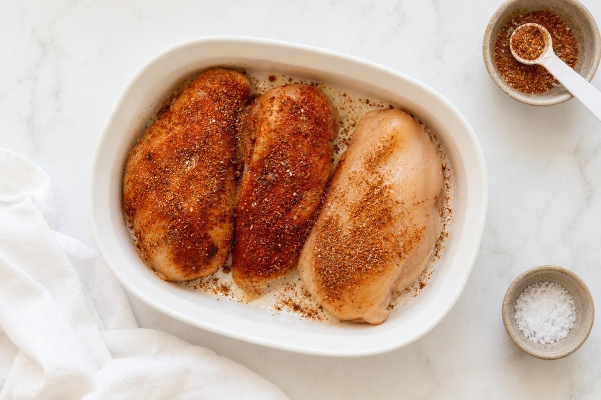 An overhead view of three raw chicken breasts in a white oval baking dish, heavily coated in a reddish spice blend, with small side bowls of extra seasoning and coarse salt nearby. An overhead view of three raw chicken breasts in a white oval baking dish, heavily coated in a reddish spice blend, with small side bowls of extra seasoning and coarse salt nearby.
