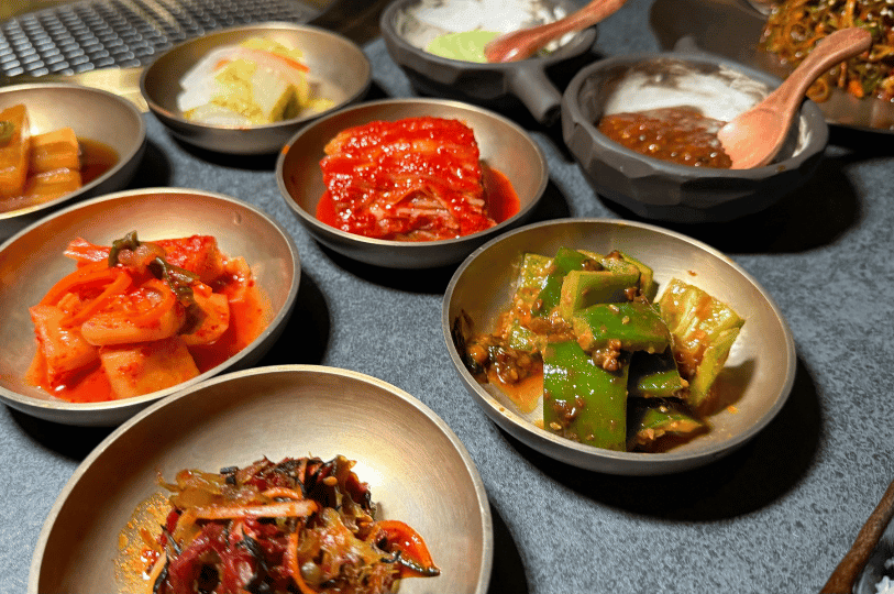 A close-up, angled view of various vibrant banchan served in traditional brass-toned bowls. The diverse textures and colors—from bright red kimchi to glossy marinated green peppers—highlight the complex flavor profiles of Korean side dishes. A close-up, angled view of various vibrant banchan served in traditional brass-toned bowls. The diverse textures and colors—from bright red kimchi to glossy marinated green peppers—highlight the complex flavor profiles of Korean side dishes.