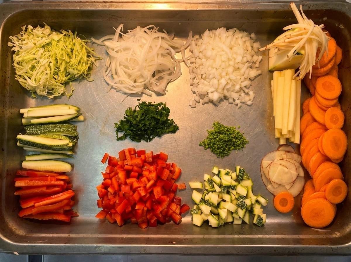 An overhead shot of a metal sheet pan organized with various prepped vegetables showcasing a variety of culinary knife cuts. An overhead shot of a metal sheet pan organized with various prepped vegetables showcasing a variety of culinary knife cuts.