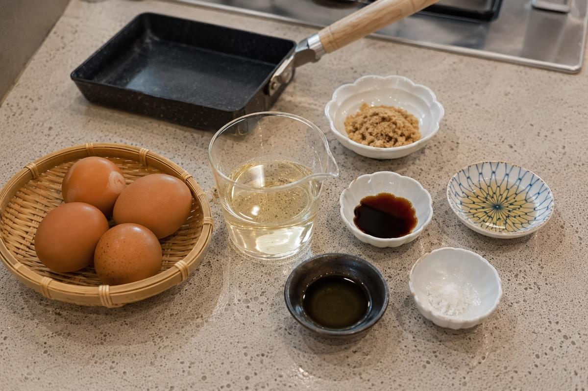An array of ingredients for tamagoyaki is neatly organized on a countertop, including a small woven basket of fresh brown eggs, a glass measuring cup of liquid dashi stock, and several small porcelain dishes containing brown sugar, soy sauce, sake, oil, and salt, positioned next to a rectangular Japanese frying pan. An array of ingredients for tamagoyaki is neatly organized on a countertop, including a small woven basket of fresh brown eggs, a glass measuring cup of liquid dashi stock, and several small porcelain dishes containing brown sugar, soy sauce, sake, oil, and salt, positioned next to a rectangular Japanese frying pan.