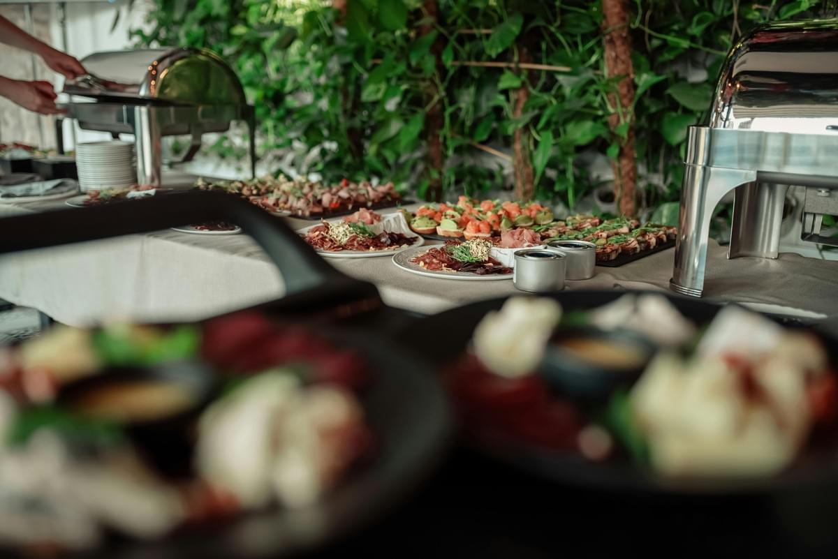 A long buffet table is spread with a colorful assortment of catered appetizers, including cured meats, crostini, and salmon bites. Stainless steel chafing dishes sit along the table set against a backdrop of dense green foliage. A long buffet table is spread with a colorful assortment of catered appetizers, including cured meats, crostini, and salmon bites. Stainless steel chafing dishes sit along the table set against a backdrop of dense green foliage.