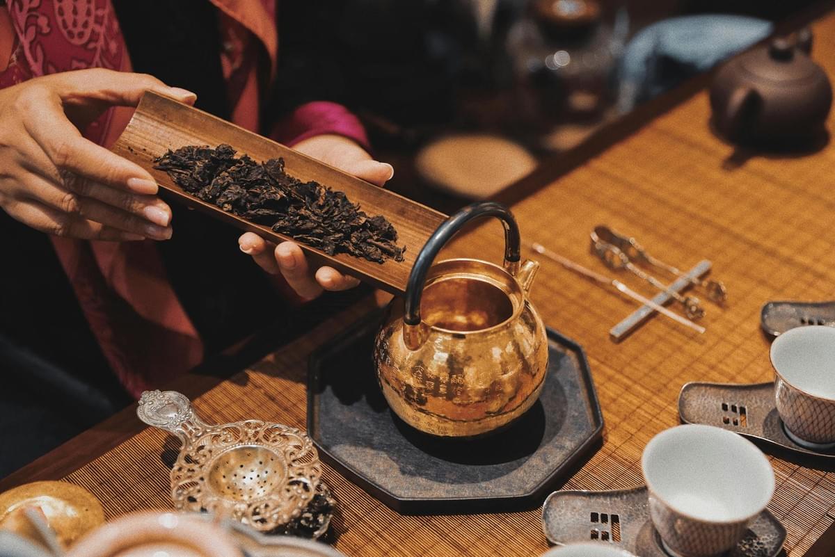 In a close-up view, a person’s hands hold a wooden scoop filled with dark, loose tea leaves, presenting them over a small, gleaming gold teapot. The scene is arranged on a textured bamboo mat, surrounded by traditional tea ceremony accessories including delicate white cups, metal tongs, and an ornate strainer. In a close-up view, a person’s hands hold a wooden scoop filled with dark, loose tea leaves, presenting them over a small, gleaming gold teapot. The scene is arranged on a textured bamboo mat, surrounded by traditional tea ceremony accessories including delicate white cups, metal tongs, and an ornate strainer.