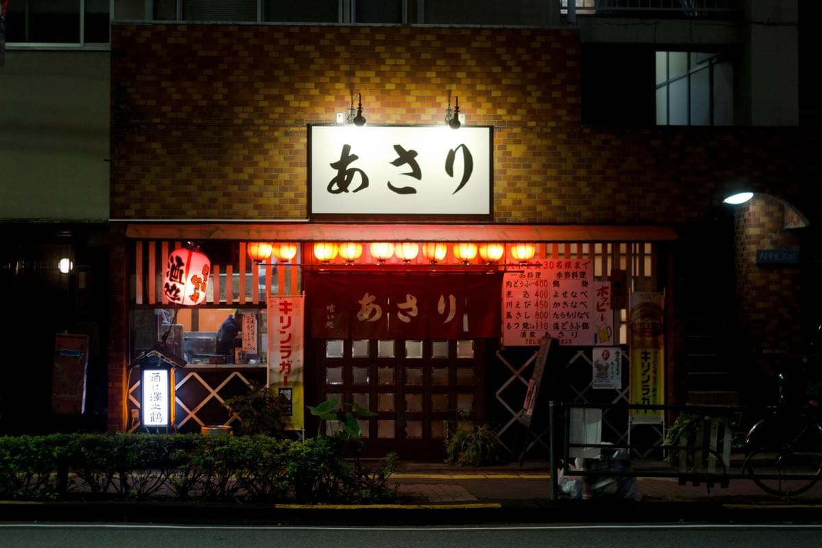 The image shows the warm, brightly lit exterior of a Japanese izakaya called "Asari" at night, featuring a brick facade and a prominent white sign. Red lanterns, a traditional noren curtain, and handwritten menu boards create a cozy, inviting atmosphere along the dark street. The image shows the warm, brightly lit exterior of a Japanese izakaya called "Asari" at night, featuring a brick facade and a prominent white sign. Red lanterns, a traditional noren curtain, and handwritten menu boards create a cozy, inviting atmosphere along the dark street.