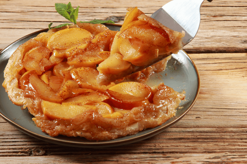 A warm slice of caramelized apple Tarte Tatin being lifted from the whole tart with a pie server on a rustic wooden table. A warm slice of caramelized apple Tarte Tatin being lifted from the whole tart with a pie server on a rustic wooden table.