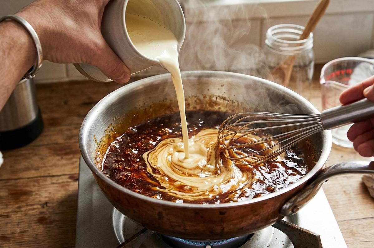 A person pouring heavy cream from a ceramic pitcher into a bubbling copper pan of dark caramel while whisking vigorously. A person pouring heavy cream from a ceramic pitcher into a bubbling copper pan of dark caramel while whisking vigorously.