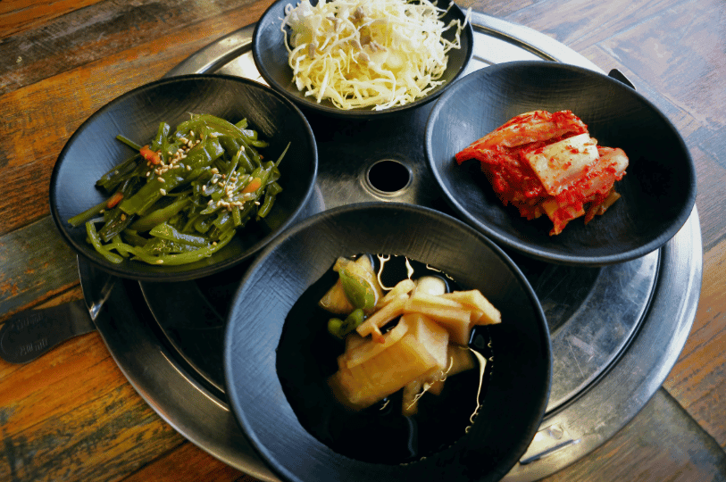 A close-up of four distinct Korean side dishes served in dark bowls arranged on a metal table grill. The selection includes shredded cabbage, seasoned seaweed stems, vibrant kimchi, and soy-pickled onions, perfectly illustrating the variety of textures and tastes in a banchan spread. A close-up of four distinct Korean side dishes served in dark bowls arranged on a metal table grill. The selection includes shredded cabbage, seasoned seaweed stems, vibrant kimchi, and soy-pickled onions, perfectly illustrating the variety of textures and tastes in a banchan spread.