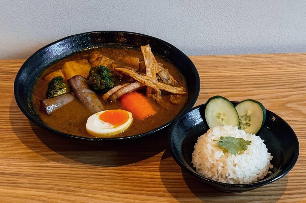 A bowl of Japanese soup curry from "Soup Curry By Ki-setsu" in Singapore, served on a light wooden tabletop alongside a side bowl of steamed white rice garnished with cilantro and sliced cucumbers, featuring rich brown broth loaded with half a soft-boiled egg, grilled eggplant, broccoli, carrots, pumpkin, and crispy burdock root strips. A bowl of Japanese soup curry from "Soup Curry By Ki-setsu" in Singapore, served on a light wooden tabletop alongside a side bowl of steamed white rice garnished with cilantro and sliced cucumbers, featuring rich brown broth loaded with half a soft-boiled egg, grilled eggplant, broccoli, carrots, pumpkin, and crispy burdock root strips.