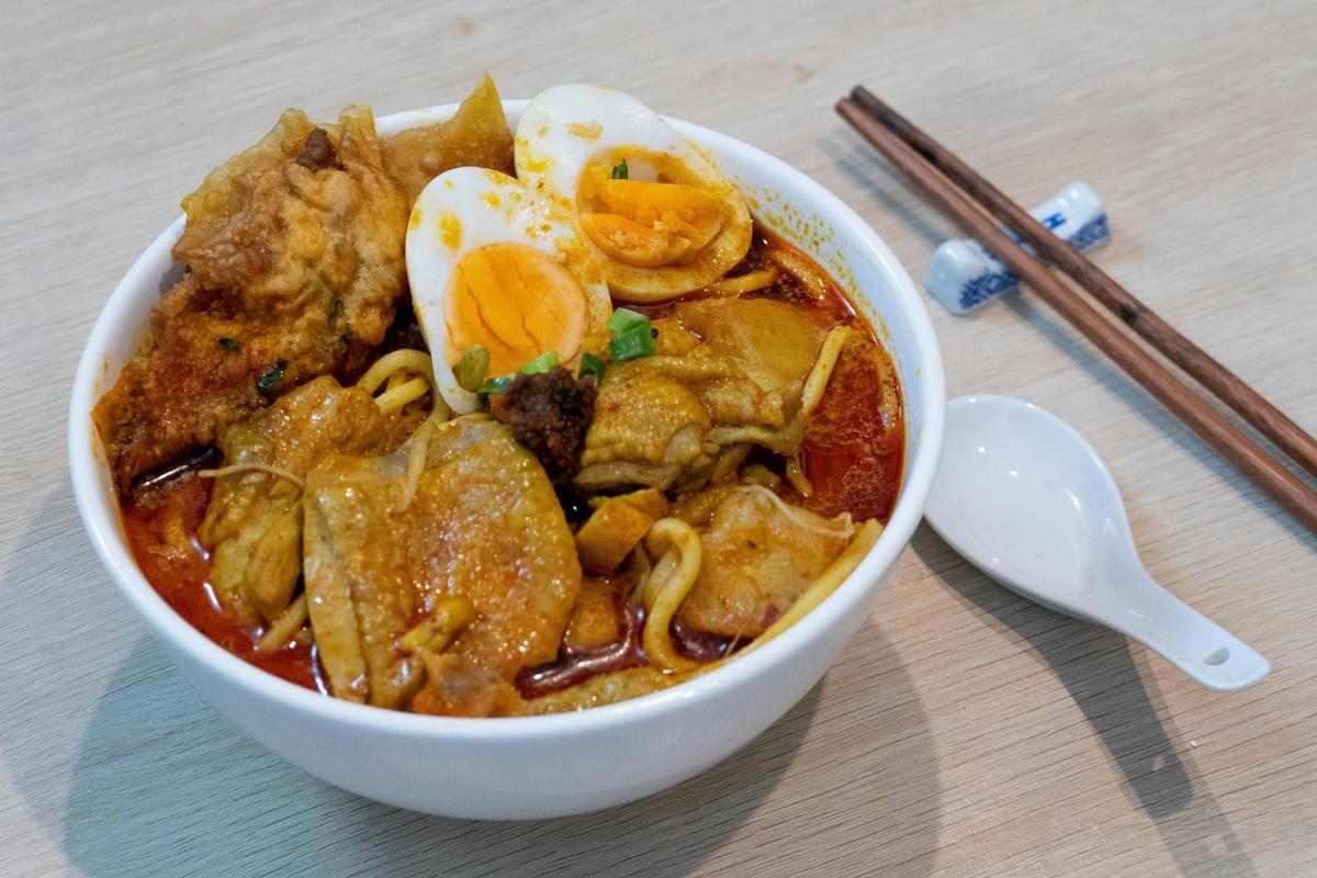 A white bowl filled with rich, spicy curry noodle soup is topped with halved hard-boiled eggs, chicken, and a crispy fried wonton. Beside the bowl on a light wooden table, a white soup spoon and wooden chopsticks rest neatly. A white bowl filled with rich, spicy curry noodle soup is topped with halved hard-boiled eggs, chicken, and a crispy fried wonton. Beside the bowl on a light wooden table, a white soup spoon and wooden chopsticks rest neatly.
