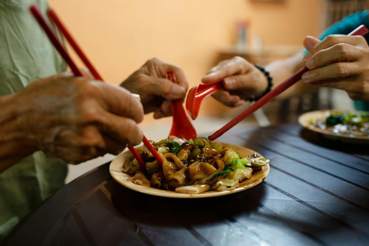 Multiple people reach with red chopsticks and spoons toward a shared plate of dark, stir-fried flat rice noodles and greens. The close-up view captures hands dining together around a dark table in a warm, casual setting. Multiple people reach with red chopsticks and spoons toward a shared plate of dark, stir-fried flat rice noodles and greens. The close-up view captures hands dining together around a dark table in a warm, casual setting.