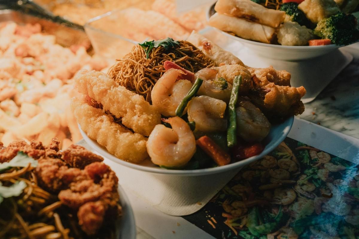 A white bowl is loaded with stir-fried noodles, sautéed shrimp, green beans, and crispy golden fried items. It sits among other buffet-style Asian dishes, including spring rolls, broccoli, and fried chicken under warm lighting. A white bowl is loaded with stir-fried noodles, sautéed shrimp, green beans, and crispy golden fried items. It sits among other buffet-style Asian dishes, including spring rolls, broccoli, and fried chicken under warm lighting.