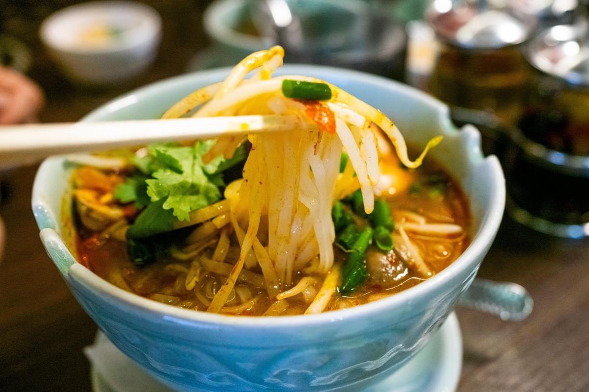 Chopsticks lift a serving of rice noodles and bean sprouts from a light blue bowl filled with flavorful broth. The dish is topped with fresh cilantro and chopped green onions in a restaurant setting. Chopsticks lift a serving of rice noodles and bean sprouts from a light blue bowl filled with flavorful broth. The dish is topped with fresh cilantro and chopped green onions in a restaurant setting.