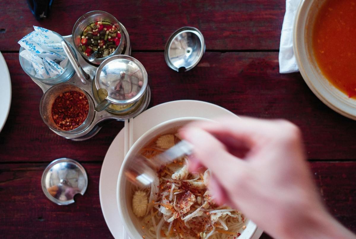 An overhead view shows a hand seasoning a bowl of noodle soup with chili flakes on a dark wooden table. A metal condiment caddy holding small glass jars of chili flakes, sliced chilis, and sugar packets sits next to the dish. An overhead view shows a hand seasoning a bowl of noodle soup with chili flakes on a dark wooden table. A metal condiment caddy holding small glass jars of chili flakes, sliced chilis, and sugar packets sits next to the dish.