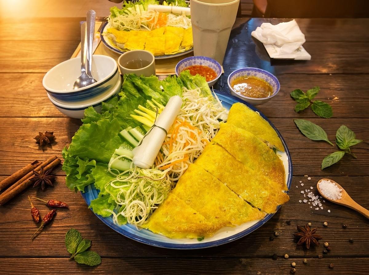 A Vietnamese bánh xèo platter on a dark wooden table, featuring sliced golden-yellow crepes next to lettuce, herbs, shredded vegetables, sprouts, and rice paper rolls, accompanied by dipping sauces, bowls, and decorative spices. A Vietnamese bánh xèo platter on a dark wooden table, featuring sliced golden-yellow crepes next to lettuce, herbs, shredded vegetables, sprouts, and rice paper rolls, accompanied by dipping sauces, bowls, and decorative spices.