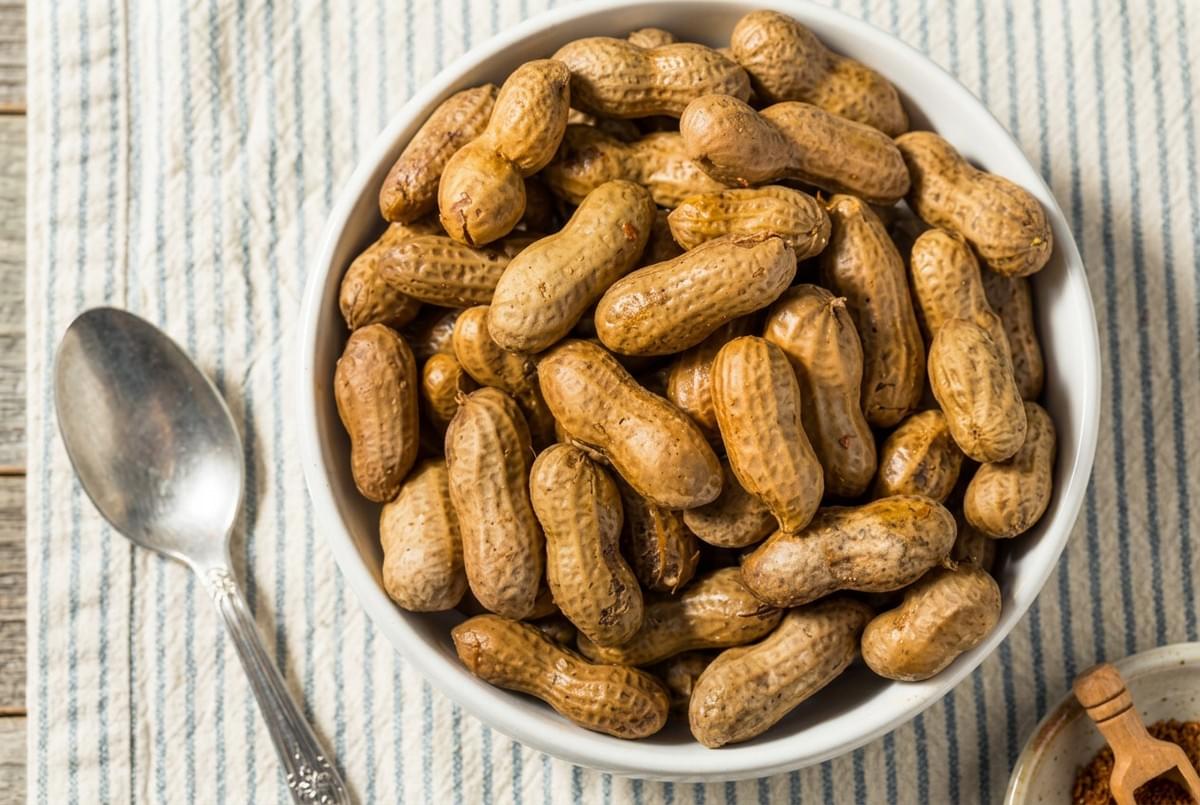 Top-down view of a white bowl filled with boiled peanuts in shells next to a spoon. Top-down view of a white bowl filled with boiled peanuts in shells next to a spoon.