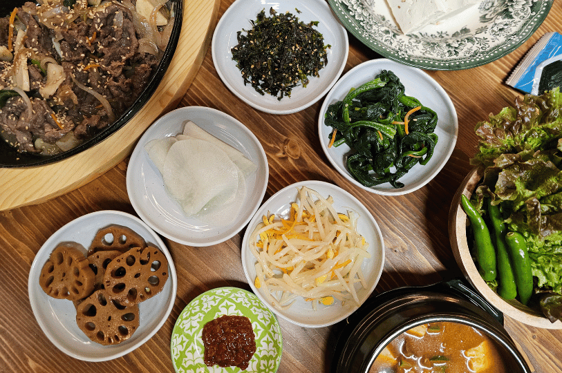 A vibrant, top-down view of a traditional Korean meal on a wooden table, showcasing the philosophy of balance with a sizzling pan of bulgogi surrounded by small white bowls of diverse banchan, including braised lotus root, seasoned spinach, bean sprouts, and fresh lettuce for wrapping. A vibrant, top-down view of a traditional Korean meal on a wooden table, showcasing the philosophy of balance with a sizzling pan of bulgogi surrounded by small white bowls of diverse banchan, including braised lotus root, seasoned spinach, bean sprouts, and fresh lettuce for wrapping.