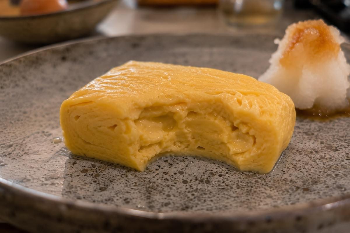 A close-up shot highlights a slice of tamagoyaki with a bite taken out, revealing its soft, layered, and juicy interior texture on a speckled grey plate next to a serving of grated daikon radish with soy sauce. A close-up shot highlights a slice of tamagoyaki with a bite taken out, revealing its soft, layered, and juicy interior texture on a speckled grey plate next to a serving of grated daikon radish with soy sauce.