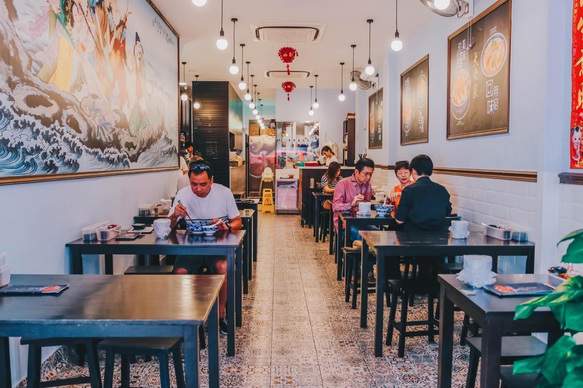 Patrons dine at dark wooden tables inside a bright, narrow restaurant with patterned floor tiles. A large Chinese artwork decorates the left wall, while framed posters and warm hanging pendant lights line the interior. Patrons dine at dark wooden tables inside a bright, narrow restaurant with patterned floor tiles. A large Chinese artwork decorates the left wall, while framed posters and warm hanging pendant lights line the interior.