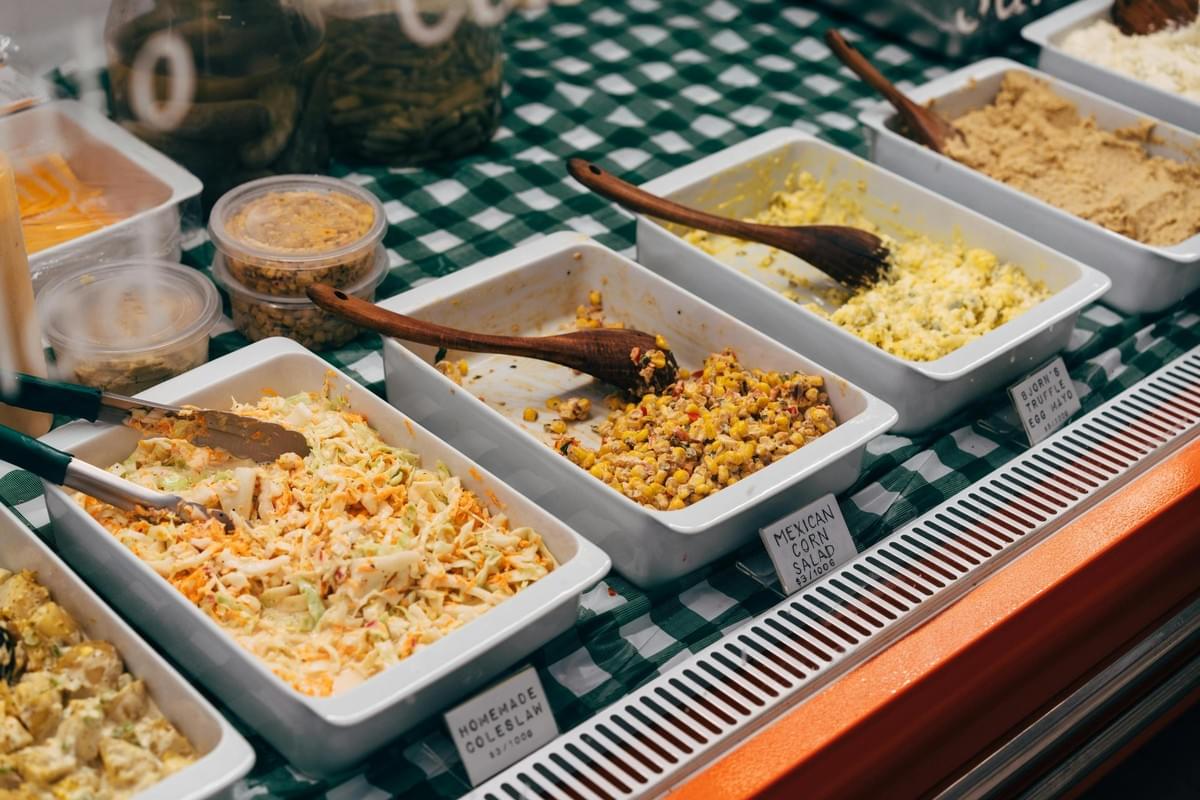 A deli display case features various prepared salads in white rectangular tubs, including coleslaw, Mexican corn salad, and truffle egg mayo. Each dish is labeled with a small handwritten price tag and served with a wooden spoon or tongs against a green checkered background. A deli display case features various prepared salads in white rectangular tubs, including coleslaw, Mexican corn salad, and truffle egg mayo. Each dish is labeled with a small handwritten price tag and served with a wooden spoon or tongs against a green checkered background.