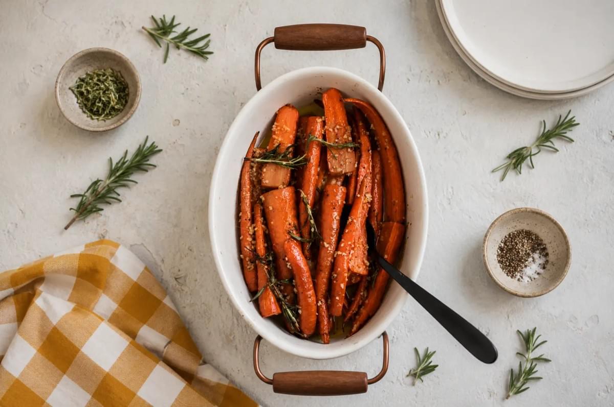 An overhead view of roasted orange carrots seasoned with garlic and rosemary sprigs inside a white oval baking dish with copper handles, surrounded by small spice bowls, fresh rosemary, and a yellow gingham cloth. An overhead view of roasted orange carrots seasoned with garlic and rosemary sprigs inside a white oval baking dish with copper handles, surrounded by small spice bowls, fresh rosemary, and a yellow gingham cloth.