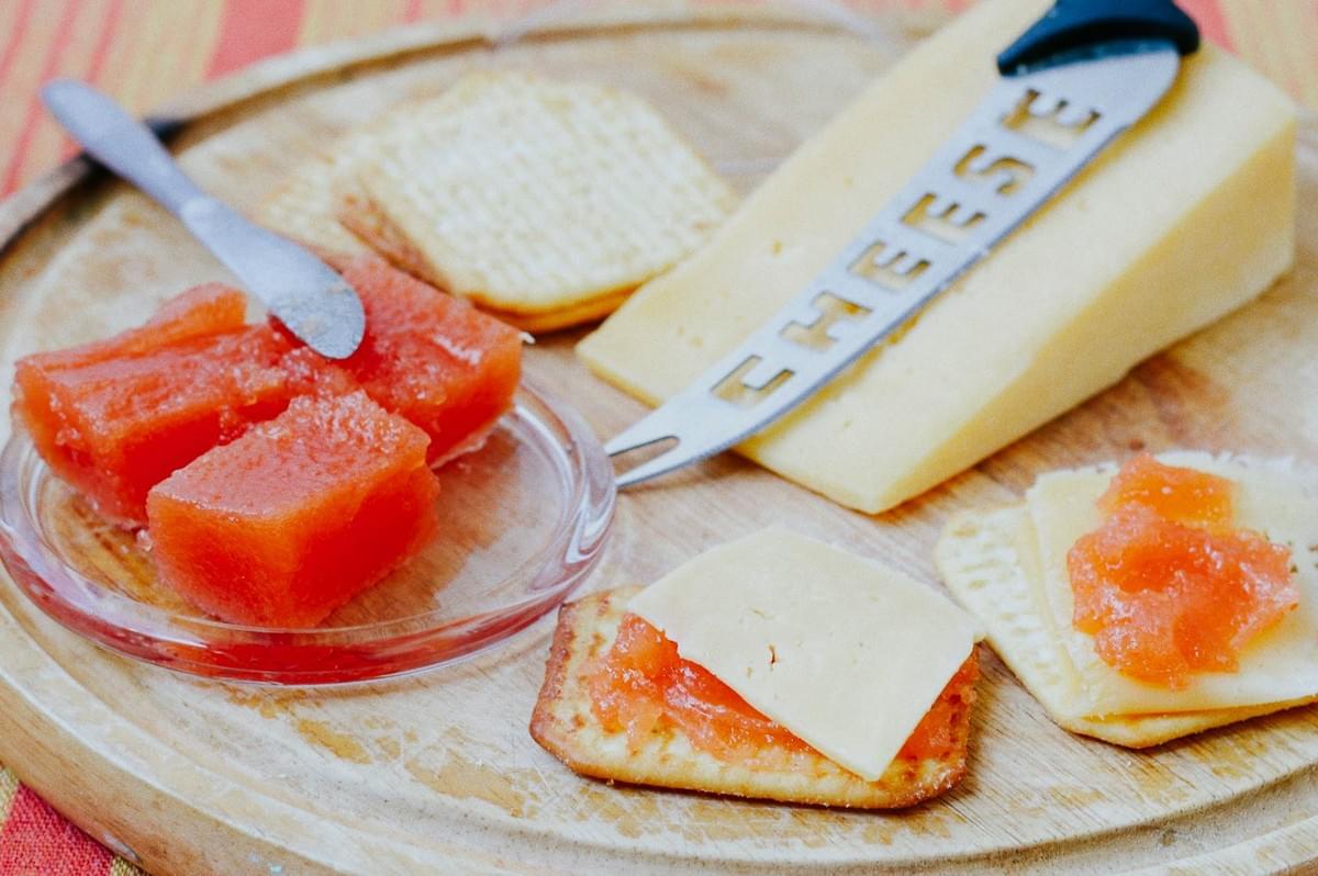 Cheese board with sliced pale yellow cheese, crackers, and quince paste cubes served with a cheese knife. Cheese board with sliced pale yellow cheese, crackers, and quince paste cubes served with a cheese knife.