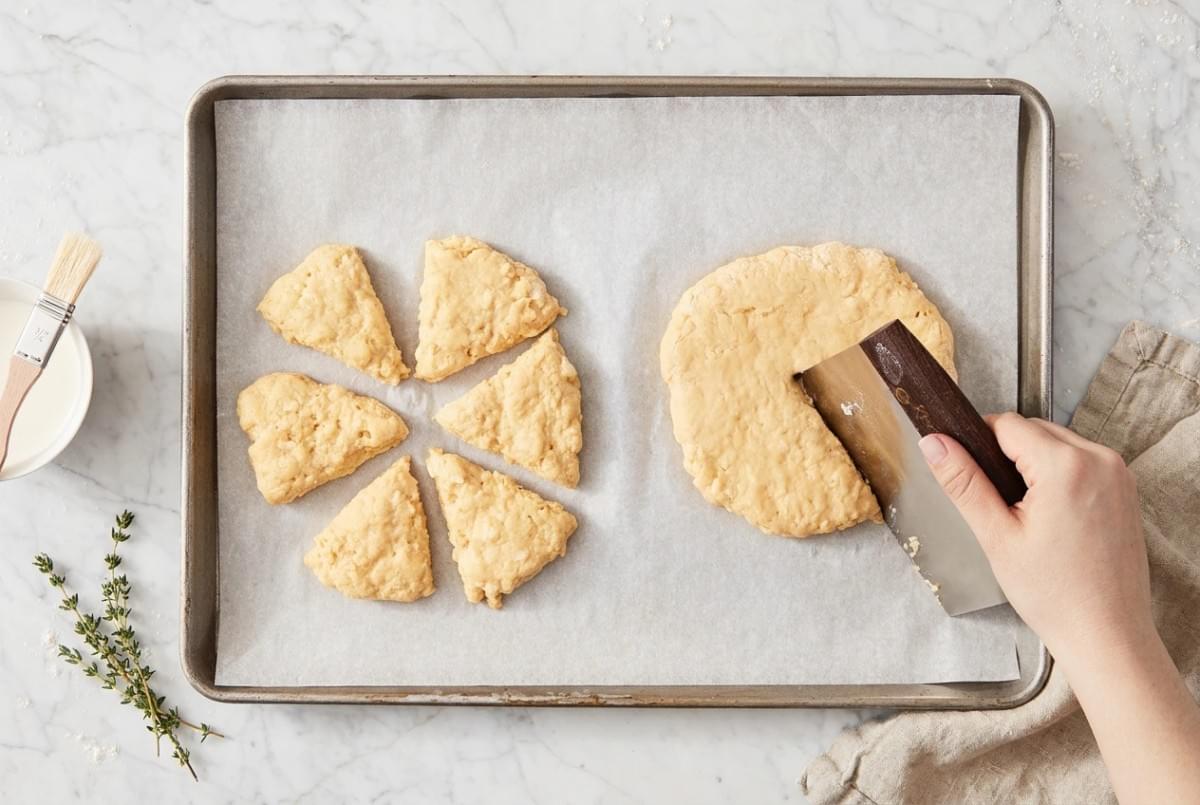 A bench scraper cutting a round of dough into triangular scone portions on a parchment-lined baking sheet. A bench scraper cutting a round of dough into triangular scone portions on a parchment-lined baking sheet.