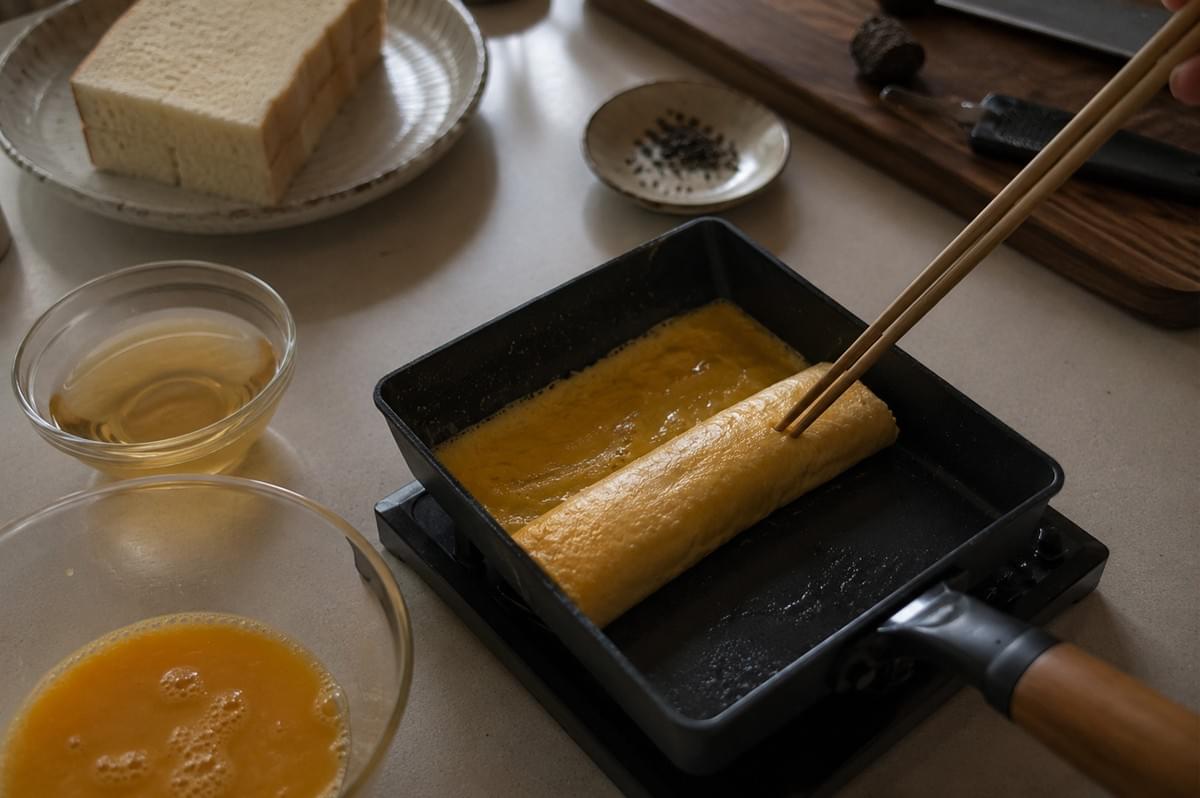 A high-angle close-up captures the meticulous process of making dashimaki tamago in a traditional square black omelet pan over a gas stove. A person uses long wooden chopsticks to carefully roll a thin, shimmering layer of cooked egg into a tight cylinder at one end of the pan, while a fresh pool of vibrant yellow beaten egg mixture coats the remaining surface. On the surrounding light-colored countertop, various preparation bowls hold dashi stock, beaten eggs, and a small plate of minced black truffles, with thick slices of white bread waiting nearby, illustrating the "patience" required for the layered cooking technique. A high-angle close-up captures the meticulous process of making dashimaki tamago in a traditional square black omelet pan over a gas stove. A person uses long wooden chopsticks to carefully roll a thin, shimmering layer of cooked egg into a tight cylinder at one end of the pan, while a fresh pool of vibrant yellow beaten egg mixture coats the remaining surface. On the surrounding light-colored countertop, various preparation bowls hold dashi stock, beaten eggs, and a small plate of minced black truffles, with thick slices of white bread waiting nearby, illustrating the "patience" required for the layered cooking technique.