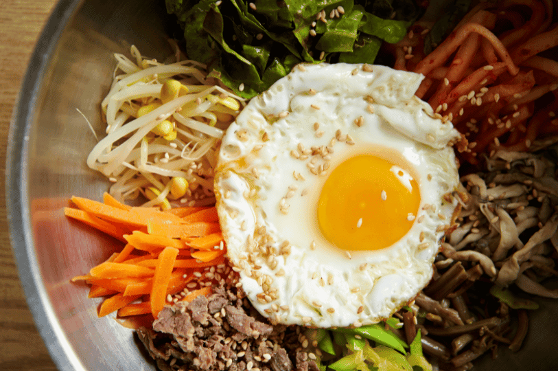 A high-angle, close-up view of a bibimbap bowl. The focus is on a fried egg sprinkled with sesame seeds, surrounded by organized portions of seasoned bean sprouts, shredded carrots, sautéed beef, and leafy greens. A high-angle, close-up view of a bibimbap bowl. The focus is on a fried egg sprinkled with sesame seeds, surrounded by organized portions of seasoned bean sprouts, shredded carrots, sautéed beef, and leafy greens.
