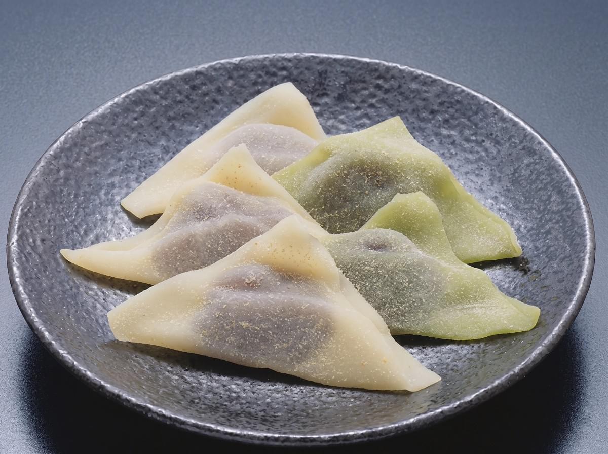 Assorted green and plain triangular yatsuhashi mochi arranged on a textured black plate. Assorted green and plain triangular yatsuhashi mochi arranged on a textured black plate.