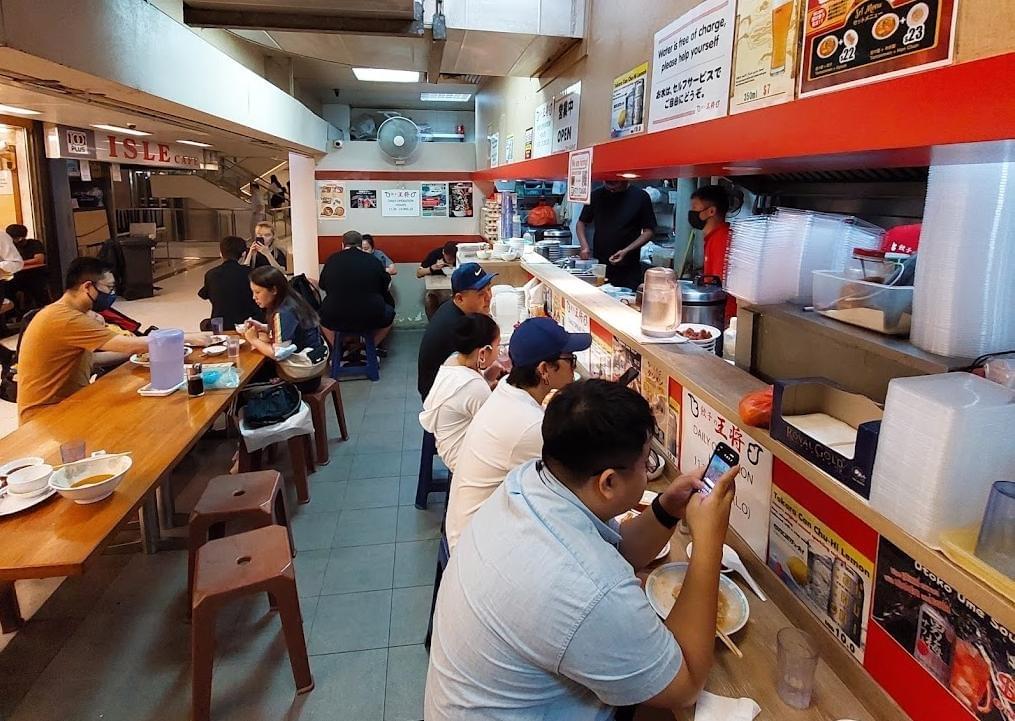Customers sit along a counter and at adjacent wooden tables inside a casual, brightly lit Asian eatery featuring an open kitchen. The restaurant has red accents, menu posters on the wall, and a glimpse of a shopping mall hallway visible in the background. Customers sit along a counter and at adjacent wooden tables inside a casual, brightly lit Asian eatery featuring an open kitchen. The restaurant has red accents, menu posters on the wall, and a glimpse of a shopping mall hallway visible in the background.