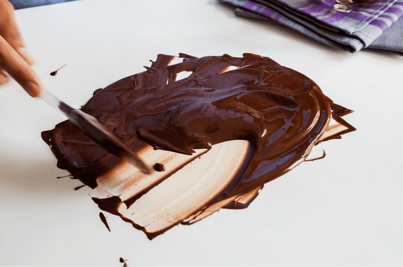A person using a metal spatula to spread and agitate melted chocolate across a smooth white marble slab to cool it during the tempering process. A person using a metal spatula to spread and agitate melted chocolate across a smooth white marble slab to cool it during the tempering process.