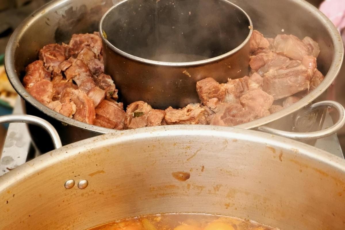 This image captures a close-up of a metal steamer insert featuring chunks of cooked meat arranged in a ring around a central chimney, with visible steam rising from the hot vessel. Beneath the meat, a large stockpot holds a simmering, reddish-brown broth, suggesting the preparation of a hearty traditional soup or stew. This image captures a close-up of a metal steamer insert featuring chunks of cooked meat arranged in a ring around a central chimney, with visible steam rising from the hot vessel. Beneath the meat, a large stockpot holds a simmering, reddish-brown broth, suggesting the preparation of a hearty traditional soup or stew.