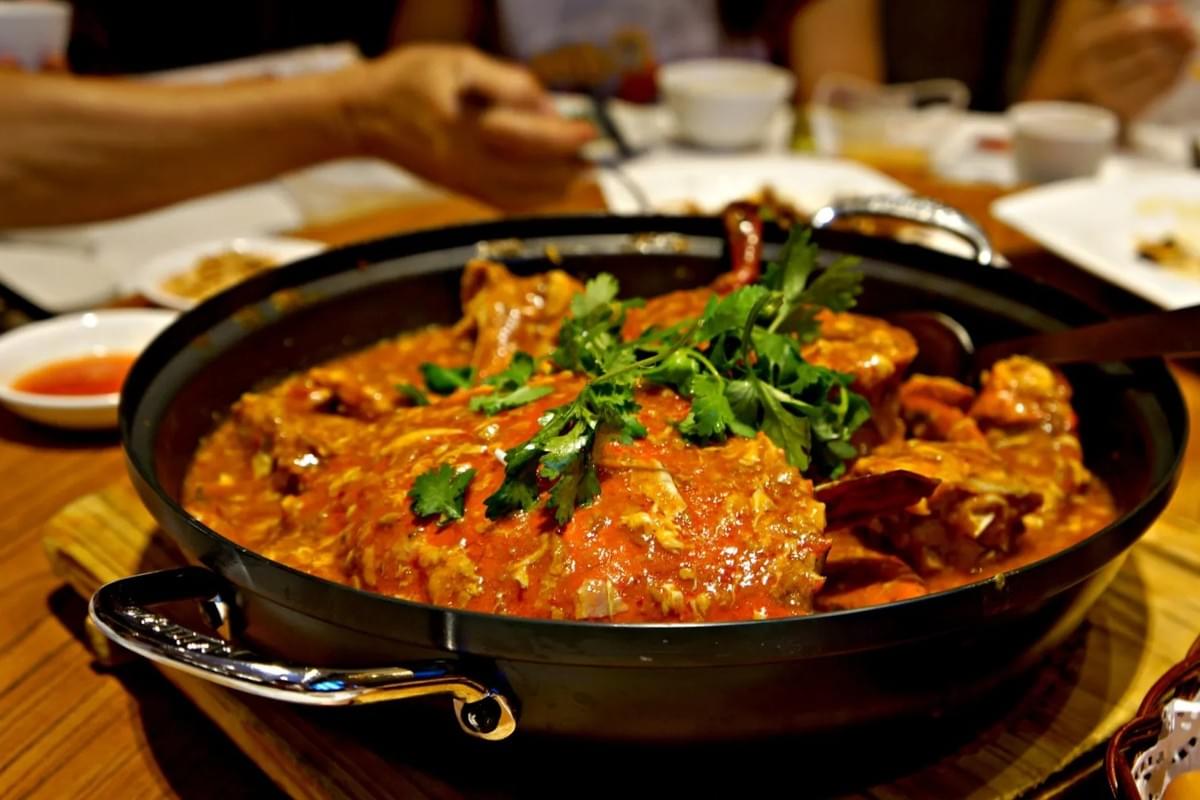 This image features a large, savory dish of chili crab served in a black pot and garnished with fresh cilantro. The crab is coated in a rich, vibrant red sauce, presented as the centerpiece of a communal dining table. This image features a large, savory dish of chili crab served in a black pot and garnished with fresh cilantro. The crab is coated in a rich, vibrant red sauce, presented as the centerpiece of a communal dining table.