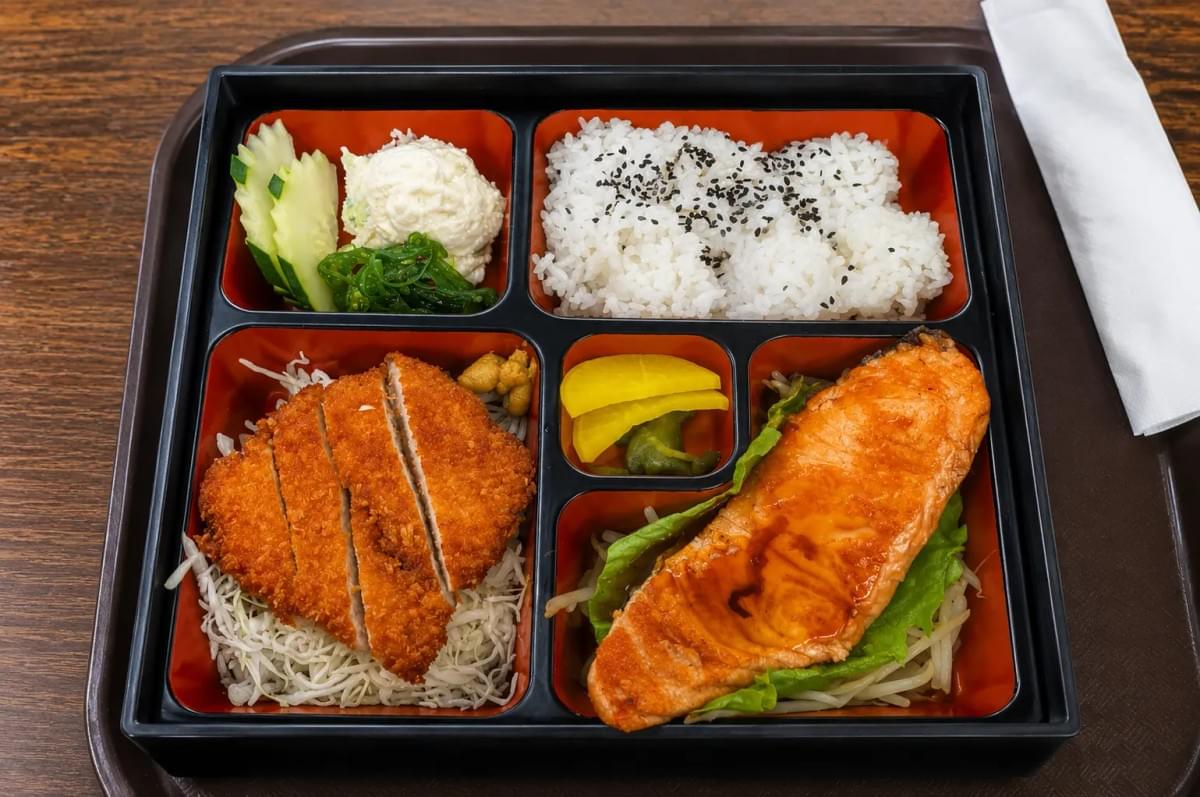 A black bento tray filled with chicken katsu, glazed salmon, sesame-seeded white rice, potato salad, seaweed, and pickled radish. A black bento tray filled with chicken katsu, glazed salmon, sesame-seeded white rice, potato salad, seaweed, and pickled radish.
