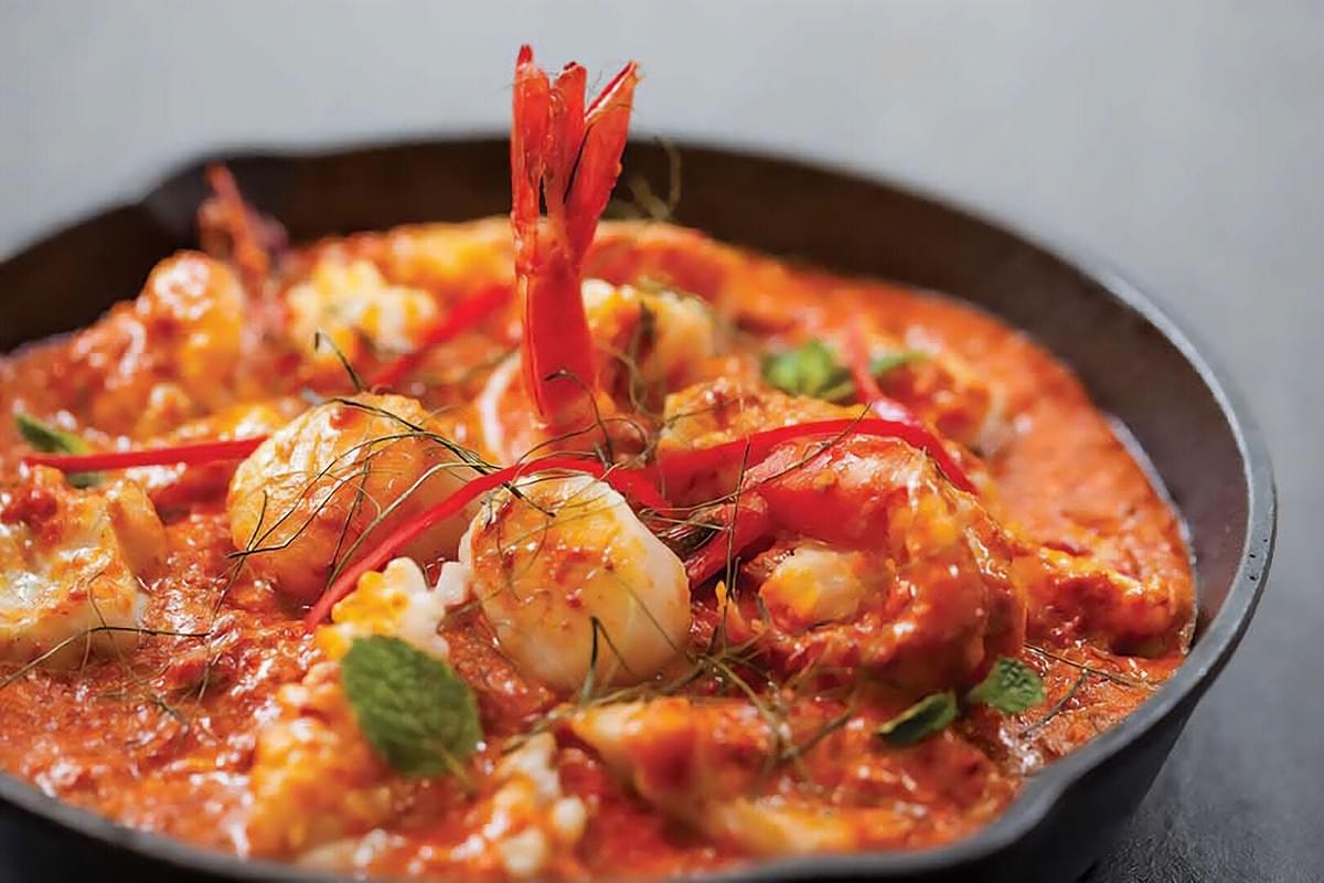 A dark skillet filled with a vibrant, spicy seafood dish in a rich orange sauce, featuring large shrimp, scallops, and squid. The dish is garnished with thin slivers of red chili pepper, julienned herbs, and fresh mint leaves. A dark skillet filled with a vibrant, spicy seafood dish in a rich orange sauce, featuring large shrimp, scallops, and squid. The dish is garnished with thin slivers of red chili pepper, julienned herbs, and fresh mint leaves.