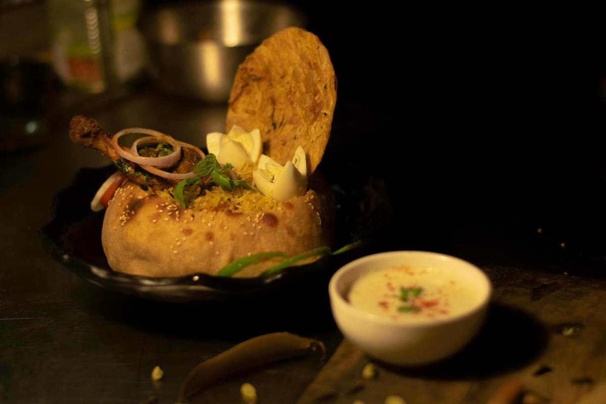 A detailed serving of biryani is elegantly presented inside a baked bread bowl, garnished with boiled eggs, sliced red onions, fresh herbs, and a roasted chicken leg. Beside the main dish sits a small white bowl of creamy raita sauce on a dark, rustic tabletop under warm lighting. A detailed serving of biryani is elegantly presented inside a baked bread bowl, garnished with boiled eggs, sliced red onions, fresh herbs, and a roasted chicken leg. Beside the main dish sits a small white bowl of creamy raita sauce on a dark, rustic tabletop under warm lighting.