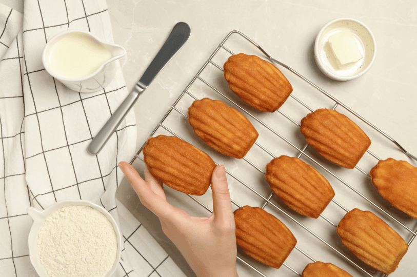 An overhead view of a hand picking up a warm madeleine from a wire rack, surrounded by baking ingredients like a bowl of flour, a pitcher of milk, and butter. An overhead view of a hand picking up a warm madeleine from a wire rack, surrounded by baking ingredients like a bowl of flour, a pitcher of milk, and butter.