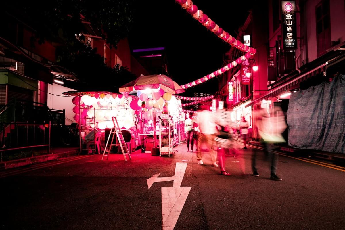 A vibrant night market street is bathed in a deep pink and red glow from overhead lanterns and illuminated stalls. Motion-blurred figures move through the scene, capturing the energetic pace of the busy urban thoroughfare. A vibrant night market street is bathed in a deep pink and red glow from overhead lanterns and illuminated stalls. Motion-blurred figures move through the scene, capturing the energetic pace of the busy urban thoroughfare.